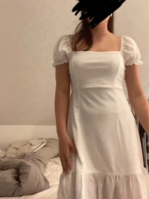 Vit puffärmad student klänning med snörning - Söt vit klänning med puffärmar och fyrkantig urringning. Klänningen har snörning i ryggen och volang nedtill, vilket ger en romantisk känsla. Tillverkad i ett lätt tyg, perfekt för varma dagar.