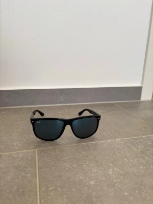 Svarta Ray-Ban Wayfarer solglasögon - Klassiska svarta Wayfarer solglasögon från Ray-Ban med bred båge och mörka glas. Snygg och tidlös design som passar till alla outfits. Ray-Ban-logga på sidan och framtill vid linsen. Perfekta för soliga dagar.