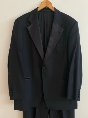 Hugo Boss smoking / smoking - Elegant svart smoking från Hugo Boss med satinrevärer, satindetaljer på fickorna och klassiska byxor med satinstripe längs sidorna. Perfekt för bröllop, gala, black tie och andra festliga tillfällen. Setet inkluderar kavaj och matchande byxor. Storlek 024 / ungefär herrstorlek M-L. Tillverkad i 100% ny ull. Begagnat i fint vintage skick med mindre bruksspår på vissa delar. Kan innehålla tecken på slitage.