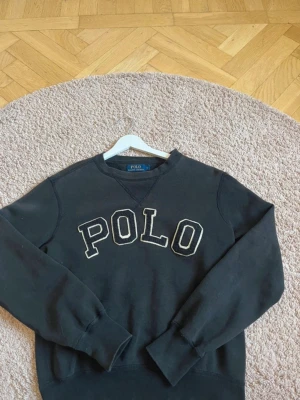 Svart sweatshirt från Polo Ralph Lauren - Svart sweatshirt från Polo Ralph Lauren med stor vit POLO-text broderad på bröstet. Klassisk rund halsringning och ribbade muddar vid ärmslut och nederkant. Perfekt för en avslappnad och stilren look.