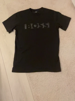 Hugo boss Tshirt  - Svart t-shirt från BOSS med stor, stilren logga i svart på bröstet. Klassisk rund halsringning och korta ärmar. Perfekt för dig som gillar en enkel men snygg look med premiumkänsla. Storlek L passar M