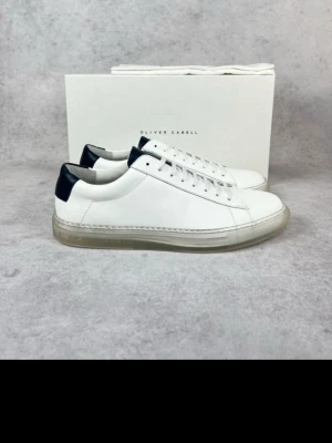 Vita sneakers från Oliver Cabell - Stilrena vita sneakers från Oliver Cabell med svart detalj på hälen och genomskinlig sula. Skorna har klassisk låg profil, vita skosnören och är tillverkade i slätt läder. Perfekta för en clean och modern look.