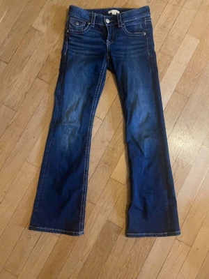 Mörkblå bootcut jeans från Gina  - Storlek 158. Snygga mörkblå bootcut jeans från Gina med klassiska fem fickor och kontrastsömmar. Jeansen har en liten ficka med lock och knapp fram samt tydliga slitningar för en cool look. Perfekta för dig som gillar en avslappnad men trendig stil.