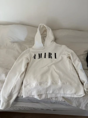 Vit hoodie från Amiri - Säljer en stilren vit hoodie från Amiri med stor svart logga över bröstet. Hoodien har klassisk huva och en stor magficka framtill. Tillverkad i mjukt bomullsmaterial som känns skönt mot huden. Perfekt för dig som gillar streetwear och vill ha en clean look.