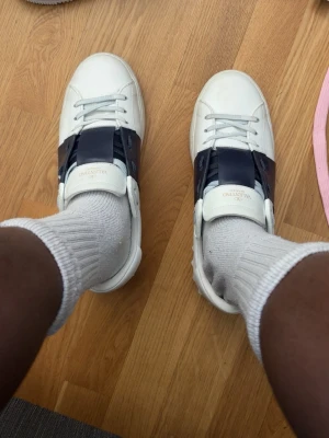 Valentino vita sneakers med mörkblå detalj - Snygga vita sneakers från Valentino med bred svart läderdetalj över mitten. Klassisk låg modell med vita skosnören och diskret logga på plösen. Perfekta för dig som gillar stilrena och exklusiva sneakers med en twist.