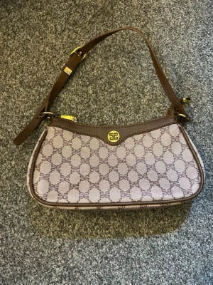 Brun axelremsväska  - Snygg axelremsväska från Givenchy i beige canvas med brunt läder och guldiga detaljer. Väskan har ett klassiskt monogrammönster med Givenchy-logga och justerbart axelband. Perfekt storlek för det viktigaste och stängs med dragkedja och tryckknapp.