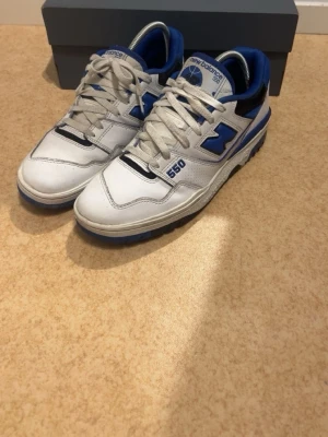 New Balance 550 vita och blå sneakers - Snygga New Balance 550 sneakers i vitt och blått med svarta detaljer. Skorna har klassisk låg siluett, perforerade paneler och tydlig branding på sidan och plösen. Ovandelen är i läder och textil, med bekväm vadderad insida och slitstark gummisula.