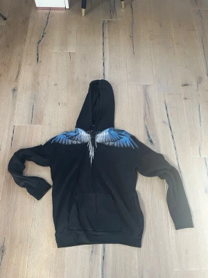 Svart hoodie med blå vingar från Marcelo Burlon - Snygg svart hoodie från Marcelo Burlon med ikoniskt vingtryck i blått och vitt över axlarna. Hoodien har huva och en stor magficka framtill. Perfekt för dig som vill sticka ut med en unik streetwear-look.