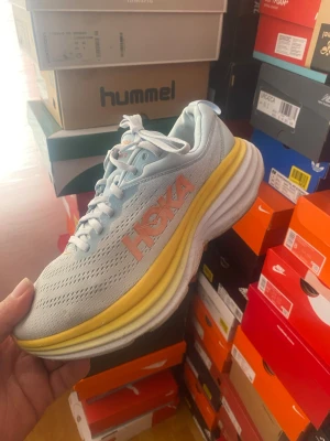 Hoka  bondi 8 vita sneakers med gul sula - Fräscha sneakers från Hoka i vitt meshmaterial med orange Hoka-logga på sidan och en tjock, gul och vit sula. Skorna har en sportig vibe och är perfekta för dig som gillar bekväma och snygga sneakers med modern design.