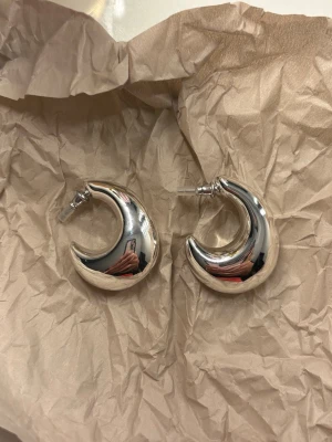 Pilgrim örhängen  - Säljer ett par chunky örhängen i silverfärgad metall med cool crescent moon-form. De har en blank, spegelblank yta och stift för hål i öronen. Perfekta för dig som gillar statement-smycken och unik design.