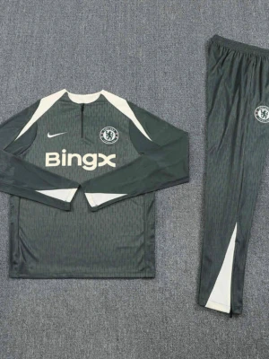 Chelsea träningsset  - Chelsea FC träningsset från Nike i mörkgrått med vita detaljer. Setet består av långärmad tröja med halv dragkedja och matchande byxor. Tröjan har Chelsea-logga och Bingx-tryck på bröstet. Tillverkat i lätt, ventilerande material perfekt för träning.