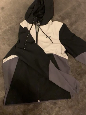 Svart, vit och grå hoodie från Under Armour - Snygg hoodie från Under Armour i svart, vitt och grått med dragkedja framtill och justerbar huva med snören. Loggan syns på bröstet och denna modellen säljs inte längre, priset kan ändras vid snabb affär tar även emot byten 