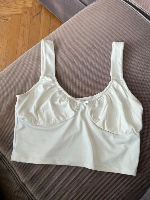 Vit mjuk bh från Divided - Söt vit bh från Divided med breda axelband och liten rosett framtill. Modellen är mjuk utan bygel och har lätt rynkade kupor för en bekväm passform. Tillverkad i ett stretchigt och mjukt material som känns skönt mot huden.