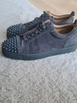 Mörkgrå sneakers med nitar - Unika mörkgrå Christian Louboutin  i mocka med coola svarta nitar över tån. Skorna har lågt skaft, svarta snören och en matchande Röd  sula som jag kan göra rent innan de skickas . Perfekta för dig som vill sticka ut med en edgy stil.Pris kan diskuteras 