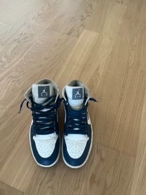 Air Jordan 1 Mid blå/vit - Säljer ett par Air Jordan 1 Mid sneakers i blått och vitt med klassisk Nike Swoosh på sidan och Air Jordan-logga vid ankeln. Skorna har högt skaft, blå snörning och perforerad tåbox. Materialet är en mix av läder och textil för en clean och sportig look.                                                                                 Pris kan diskuteras!🤝🔥