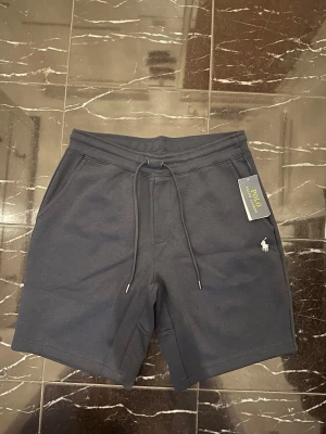 Mörkblå shorts från Polo Ralph Lauren - Mörkblå shorts från Polo Ralph Lauren med snörning i midjan och klassisk logga broderad framtill. Tillverkade i mjuk bomull, har sidofickor och en bakficka. Perfekta för en chill och stilren look.