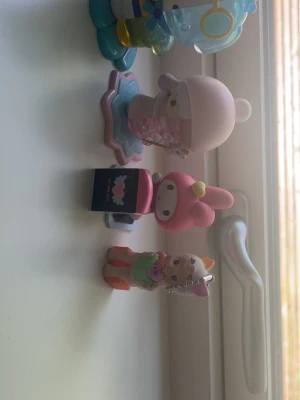 Söta Sanrio-figurer och nyckelring - Samling med fyra Sanrio-inspirerade figurer i plast, inklusive My Melody och en liten figur med trollstav på stjärnformad bas. Färgerna är mestadels rosa, blå, vitt och lite orange. En figur är en nyckelring med kattöron och klänning. Perfekt för dekoration eller samling.