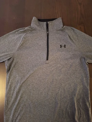 Under Armour träningströja - Snygg grå långärmad träningströja från Under Armour med halv dragkedja och hög krage. Tillverkad i ett mjukt och stretchigt funktionsmaterial som passar perfekt till träning eller löpning. Diskret logga på bröstet och svart insida på kragen.