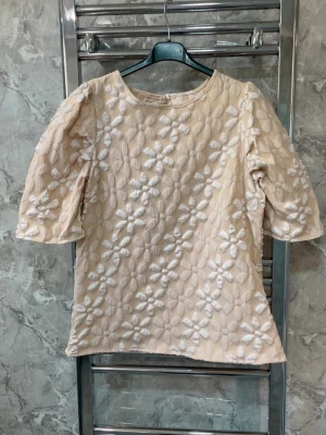 Beige blus med vita broderade blommor - Supersöt beige blus med puffiga halvlånga ärmar och vita broderade blommor över hela plagget. Blusen har rund halsringning och stängs med knapp i nacken. Perfekt för dig som gillar romantiska detaljer och unik struktur.