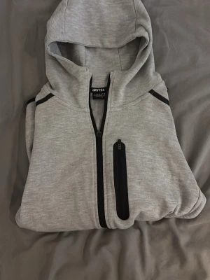 Ljusgrå hoodie från Drytex - Snygg ljusgrå hoodie från drytex med hel dragkedja framtill och svarta detaljer vid dragkedjor och axlar. Hoodien har en bröstficka med dragkedja och en klassisk huva. Tillverkad i mjukt och bekvämt material, perfekt för chill dagar.