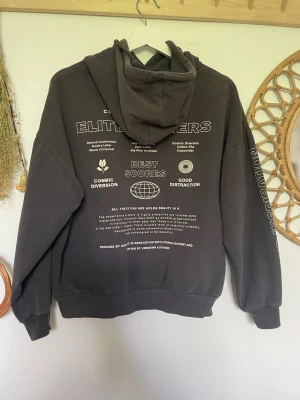 Mörkgrå hoodie Elite Winners - Mörkgrå hoodie med tryck både fram och bak samt på ena ärmen. På bröstet finns en palm och texten 'Elite Winners'. Baksidan har flera grafiska tryck och texter.