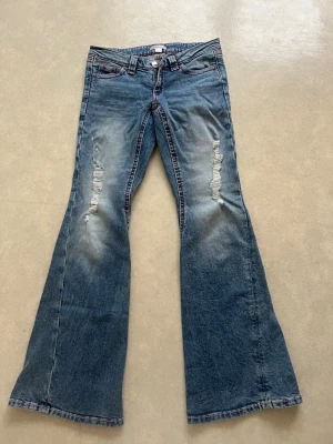 Blå bootcut jeans med slitningar - Säljer ett par blå bootcut jeans med slitningar på framsidan och broderade detaljer på bakfickorna. Jeansen har låg midja och klassisk femficksdesign. Materialet är denim i bomull och passformen är utsvängd nedtill för en retro vibe.