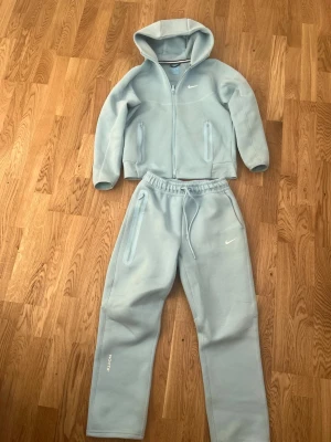 Noctatech ljusblå  - Säljer ett ljusblått loungewear set från Nike NOCTA med hoodie och matchande byxor. Setet har dragkedjor på både jacka och byxor, samt diskreta loggor på bröstet och benet. Materialet är mjukt och bekvämt, perfekt för chill dagar hemma.
