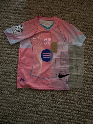 Rosa FC Barcelona matchtröja Nike - Säljer en rosa och ljusblå FC Barcelona matchtröja från Nike med klubbmärke, Spotify-logga och Champions League-märke på ärmen. Tröjan har korta ärmar, V-ringning och är tillverkad i ett lätt, ventilerande material perfekt för fotboll.