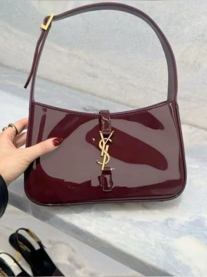 Vinröd YSL handväska i lack - Snygg vinröd handväska från Saint Laurent i glansigt lackmaterial. Väskan har en klassisk axelrem, guldigt YSL-logo framtill och en stilren, rektangulär form. Perfekt accessoar för att lyfta din outfit med en lyxig touch.