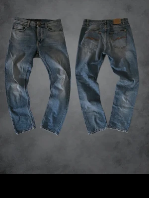 Blå jeans från Nudie Jeans - Säljer ett par klassiska blå jeans från Nudie Jeans med snygga slitningar och kontrastsömmar. Jeansen har fem fickor, rak passform och läderpatch med logga bak. Perfekta för dig som gillar en avslappnad och tidlös look.