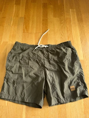 Olivgröna/militärgröna badbyxor från Urban Classics - Säljer ett par olivgröna/militärgröna badbyxor från Urban Classics med elastisk midja och vit dragsko. Klassisk modell med kortare ben och en liten logotyp-patch framtill. Materialet är snabbtorkande och lätt, perfekt för stranden eller poolen.