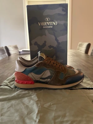 Valentino Rock Runners  - Rock Runners från Valentino Garavani med camomönster i grönt, blått och beige. Skorna har bruna mockadetaljer, röda nitar vid hälen och vita samt svarta inslag. Klassisk låg modell med bruna skosnören, perfekt för både vardag och finare tillfällen. Jättebra skick och säljer då jag har växt ur dom. Storleken är 41 men passar även 42. Hör av er vid funderingar. Kom gärna med bud😁