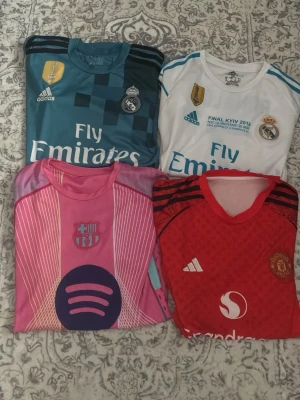 Real Madrid, Manchester United och Barcelona matchtröjor - Fyra snygga fotbollströjor: två Real Madrid-tröjor i vitt och turkos med guld- och blå detaljer, en rosa Barcelona-tröja med Spotify-logga och en röd Manchester United-tröja. Alla är kortärmade och perfekta för fotboll eller träning. Stygn har lossnat på något ställe men det syns inte när man har på sig dem. De är inte äkta men de är bra kvalite
