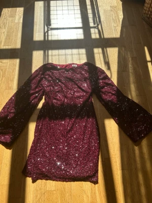 Vinröd glittrig paljettklänning - Säljer en vinröd kortklänning täckt av glittriga paljetter. Klänningen har långa ärmar och en figurnära passform som ger en snygg siluett. Perfekt för dig som vill sticka ut med extra mycket glitter och glam.