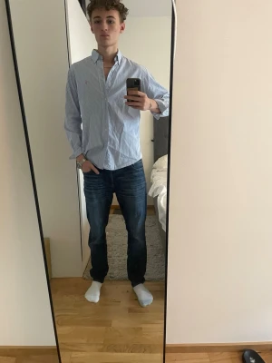 Blårandig skjorta från Ralph Lauren - Klassisk blårandig skjorta från Ralph Lauren med button-down krage och diskret rosa logga på bröstet. Skjortan har lång ärm och knäppning framtill. Perfekt för en clean och stilren look.