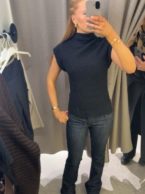 Svart ribbad polotopp - Snygg svart ribbad polotopp med hög hals och ärmlös design. Toppen har en tight passform och är perfekt att styla med jeans eller kjol. Materialet är stretchigt och bekvämt, vilket gör den enkel att bära hela dagen.