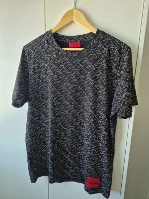Hugo t-shirt  - Snygg svart t-shirt från Hugo med vitt grafiskt mönster över hela tröjan och en röd Hugo-label nertill. Klassisk rund hals och korta ärmar. Perfekt för dig som gillar streetstyle och vill sticka ut lite extra.
