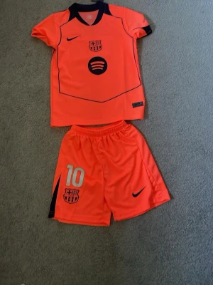 Barça träningsset Nike orange - Säljer ett orange FC Barcelona träningsset från Nike med klubbmärke och Spotify-logga på tröjan samt nummer 10 på shortsen. Setet består av en kortärmad tröja och shorts i lätt och ventilerande material, perfekt för fotbollsträning.