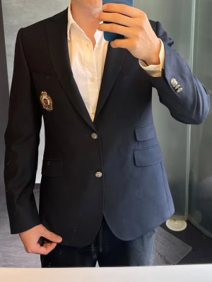 Mörkblå Morris kavaj  - Stilren mörkblå kavaj från Morris med två knappar framtill och ett emblem på bröstet. Klassisk passform med två fickor med lock och en bröstficka. Perfekt för en dressad look. Den har storlek 48 vilket motsvarar storlek M 