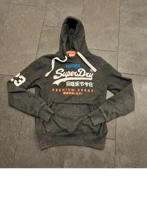 Mörkgrå Superdry hoodie med tryck - Snygg mörkgrå hoodie från Superdry med stort vitt och orange tryck på bröstet och siffran 23 på ärmen. Klassisk känguruficka, huva med vita snören och mjukt material. Perfekt för en avslappnad streetstyle-look. Pris kan diskuteras. MVH stentorparchive⚓️
