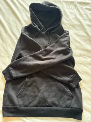 Svart hoodie med magficka - En klassisk svart hoodie med huva och dragsnören. Hoodien har en stor magficka framtill och ribbade muddar vid ärmslut och nederkant. Perfekt för en avslappnad och stilren look. Materialet känns mjukt och bekvämt, troligen bomullsblandning.
