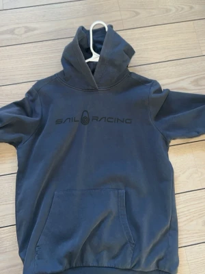 Mörkblå hoodie från Sail Racing - Mörkblå hoodie från Sail Racing med stor ficka framtill och logga tryckt över bröstet. Klassisk huva och lång ärm, perfekt för en avslappnad stil. Tillverkad i mjuk bomull för extra komfort.