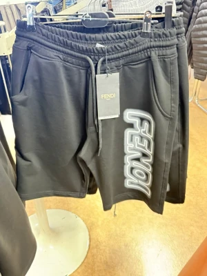 Svarta shorts från Fendi med tryck - Svarta shorts från Fendi med stort vitt och grått tryck på ena benet. De har elastisk midja med snörning och två sidofickor. Perfekta för en avslappnad och trendig stil.