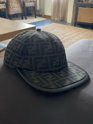 Svart keps från Fendi med FF-mönster - Säljer en svart keps från Fendi med det klassiska FF-logomönstret över hela kepsen. Skärmen har en snygg kant i svart läderimitation som ger en exklusiv känsla. Perfekt accessoar för dig som gillar lyxiga detaljer och streetstyle. DEN ÄR SPRILLANS NY!