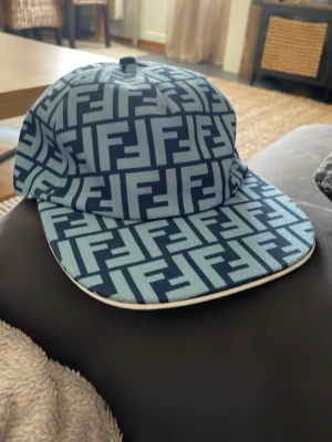 Blå Fendi keps med FF-mönster - Snygg blå keps från Fendi med ikoniskt FF-logomönster över hela kepsen. Kepsen har en rak skärm och är tillverkad i ett slitstarkt tygmaterial. Perfekt för dig som vill sticka ut med en trendig accessoar. SPRILLANS NY!