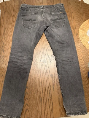 Nudie Jeans Lean Dean Mörkgrå - Säljer ett par grå jeans från Nudie Jeans med klassisk femficksmodell och raka ben. Jeansen har en diskret tvättad look och Nudie Jeans signaturbrodyr på bakfickorna. Materialet är mjukt denim i bomull som ger en skön känsla.