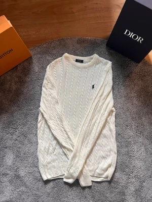 Vit kabelstickad tröja Polo Ralph Lauren - Kabelstickad vit tröja från Polo Ralph Lauren med rund halsringning och den klassiska broderade loggan på bröstet. Tröjan har långa ärmar och ribbade muddar vid ärmslut och nederkant. Perfekt för en clean och stilren look.