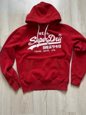 Röd Superdry hoodie med tryck -  Priset är inte fast i sten.                                             Röd hoodie från Superdry med vit logga och japansk text på bröstet. Klassisk känguruficka framtill, vita dragsnören i huvan och ribbade muddar. Perfekt för en avslappnad streetstyle-look.