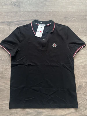 Svart Moncler pikétröja med röd/vit detalj - Snygg svart pikétröja från Moncler med klassisk krage och knappar framtill. Tröjan har röda och vita ränder på krage och ärmslut samt Moncler-logga broderad på bröstet. Perfekt för en stilren och sportig look.