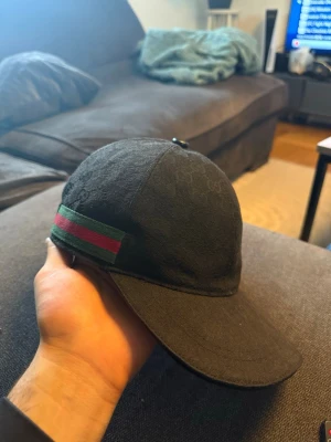 Svart keps från Gucci med GG-mönster - Snygg svart keps från Gucci med det klassiska GG-monogrammönstret över hela kepsen. På sidan finns en grön och röd rand som ger den där ikoniska Gucci-looken. Justerbar rem bak i läder och kepsen är tillverkad i ett slitstarkt tyg.